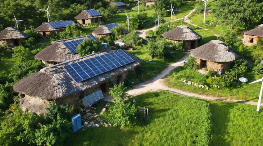 "Aerial view of an off-grid community with solar panels and wind turbines, highlighting the integration of renewable energy systems within a green, self-sufficient environment."