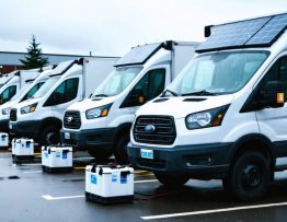 Portable modular solar panels and rugged battery cases charging a row of evacuation vans at a municipal staging lot under bright overcast skies, with wet pavement and emergency staff working in the background.