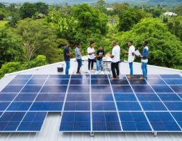 "Community members of diverse backgrounds engaged in a planning meeting with financial advisors and technical experts, discussing the implementation of solar microgrids surrounded by rooftops covered with solar panels and lush greenery, symbolizing the fusion of sustainable energy and community-driven finance."