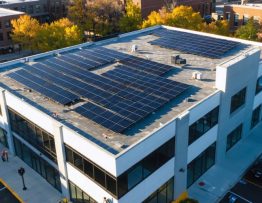 Aerial 3/4 view of a mid-rise office building with a large rooftop solar panel array at golden hour, city skyline and EV chargers visible in the background.
