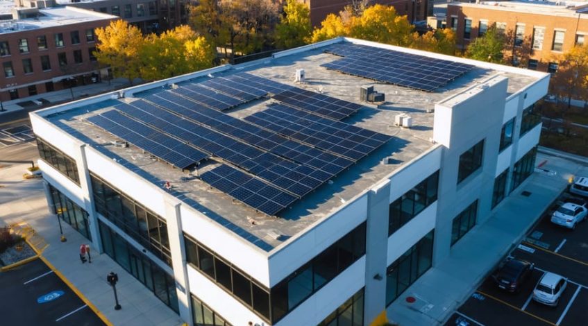 Aerial 3/4 view of a mid-rise office building with a large rooftop solar panel array at golden hour, city skyline and EV chargers visible in the background.