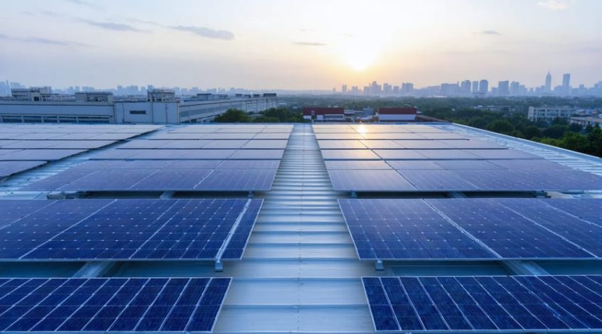 Elevated wide view of blue solar panels covering a commercial warehouse roof at golden hour, with blurred city skyline, loading bays, and rooftop HVAC units in the background.