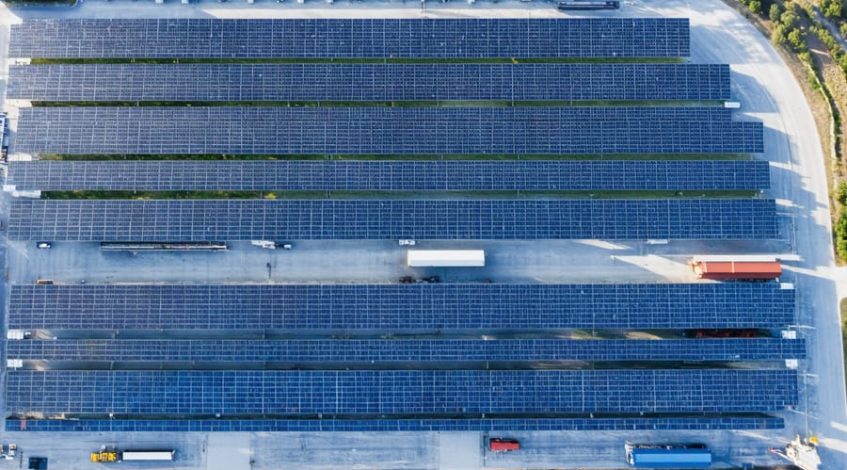 Aerial oblique view of a community solar farm next to a modern distribution center with unbranded trucks, lit by warm sunrise, illustrating commercial access to off-site renewable energy.