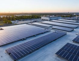 A 45-degree aerial view of a commercial facility with rooftop solar panels and adjacent battery storage units at golden hour, with distant transmission lines and a soft city skyline in the background.