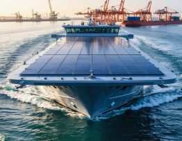 Modern cargo-ferry hybrid ship with large solar panels across its upper deck, photographed from above the starboard bow at golden hour, with blurred port cranes and coastline in the background over calm blue water.