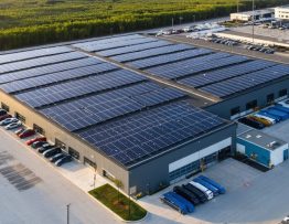 Oblique aerial view of a commercial building with rooftop solar panels and containerized battery storage, with distant transmission lines and a faint city skyline at golden hour.