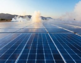 Ground-level view of a commercial solar farm with long rows of blue panels and a gravel firebreak, lit by smoky golden-hour sunlight, with a distant wildfire line and rising smoke over scrub-covered hills.