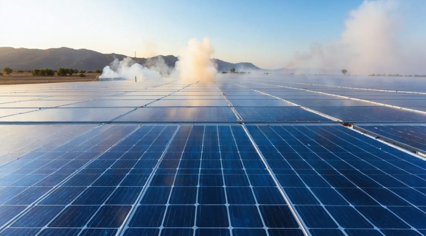 Ground-level view of a commercial solar farm with long rows of blue panels and a gravel firebreak, lit by smoky golden-hour sunlight, with a distant wildfire line and rising smoke over scrub-covered hills.