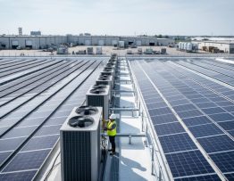 Hard-hat engineer inspecting solar panels on a commercial rooftop near HVAC units under bright overcast light, with industrial skyline and loading docks in the background.