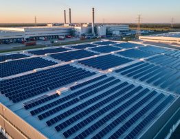 Aerial oblique view of a modern manufacturing plant with rows of blue solar panels covering the rooftop at golden hour, with factory buildings, smokeless chimneys, loading docks, and distant power lines in the background.
