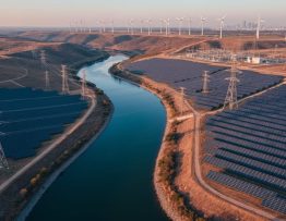 Elevated panoramic view of a river acting as a boundary with large solar farms on both banks, linked by high-voltage lines, with wind turbines, a substation, and a faint city skyline under warm evening light.