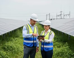 Two professionals in safety vests and hard hats review a tablet beside a large community solar farm, with rows of ground-mounted panels and distant utility poles under soft overcast daylight.