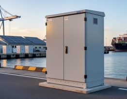 Weatherproof microgrid interconnection switchgear cabinet at a solar-powered port with cranes, rooftop solar panels, battery storage containers, and a moored cargo ship in warm early light