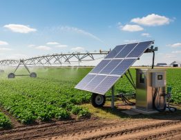 Ground-mounted solar panels beside an electric irrigation pump feeding a center-pivot sprinkler system across a large green crop field under bright daylight, with distant barns and silos in the background.