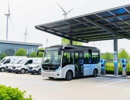 Electric shuttle bus and delivery vans charging at a renewable-powered depot with solar panels overhead and a clean modern facility in the background.