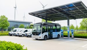 Electric shuttle bus and delivery vans charging at a renewable-powered depot with solar panels overhead and a clean modern facility in the background.